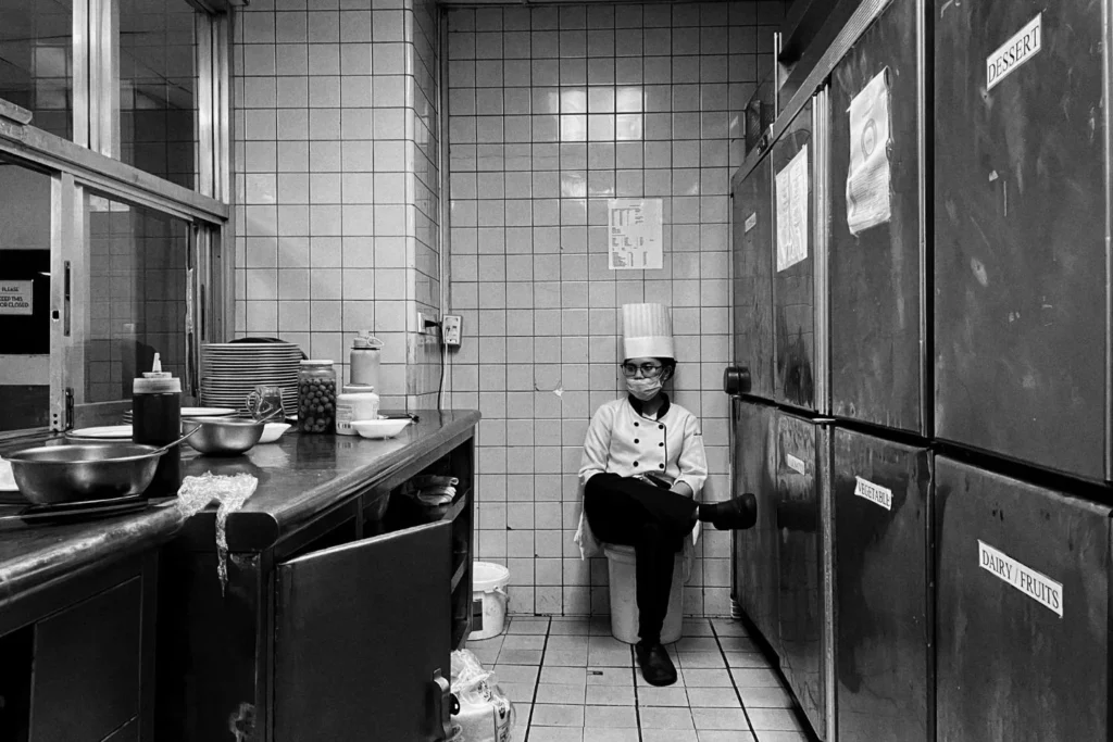 A black and white wide shot of a female chef in full uniform and face mask, sitting on a bucket in a narrow kitchen hallway between large industrial refrigerators labeled 'Dessert' and 'Vegetable'.