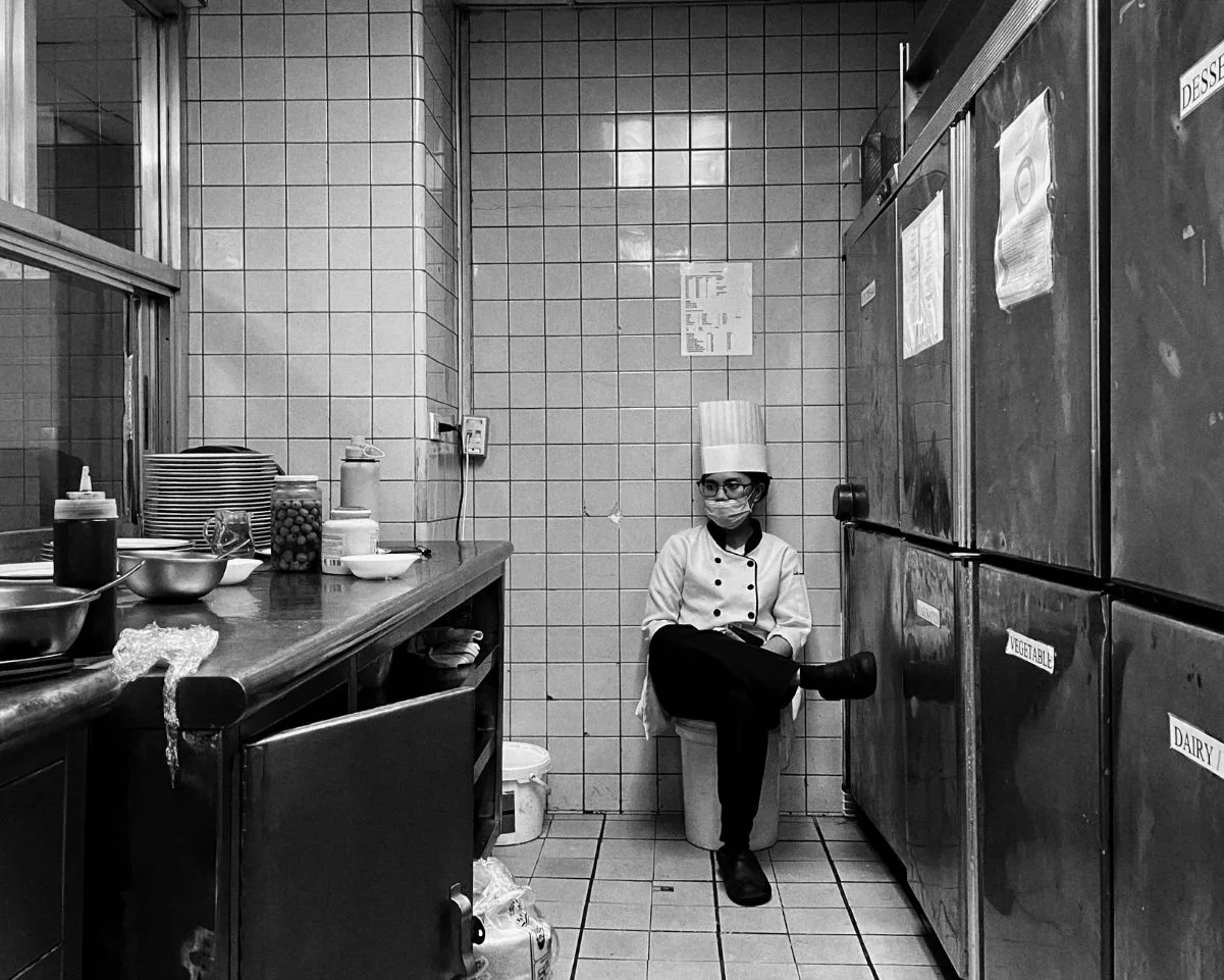 A black and white wide shot of a female chef in full uniform and face mask, sitting on a bucket in a narrow kitchen hallway between large industrial refrigerators labeled 'Dessert' and 'Vegetable'.