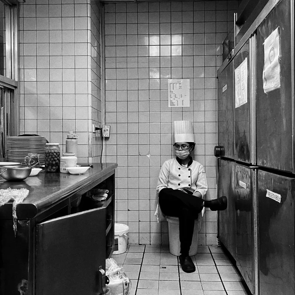 Powerful black and white documentary-style photograph of a weary young chef, fully dressed in chef whites and a tall hat, sitting alone on a bucket in a cluttered storage corridor, reflecting the physical and emotional exhaustion within a toxic kitchen culture.