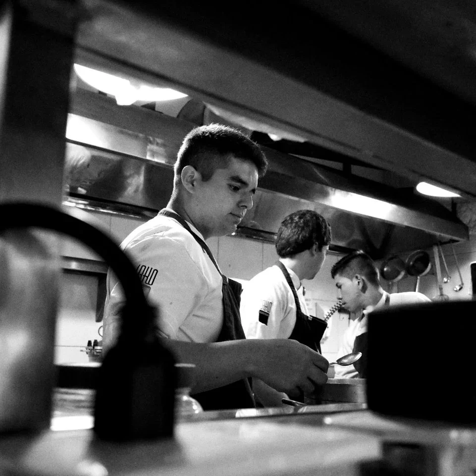 Black and white high-contrast photo of chefs working on a kitchen line, illustrating the intense environment and physical demands of restaurant industry labor.