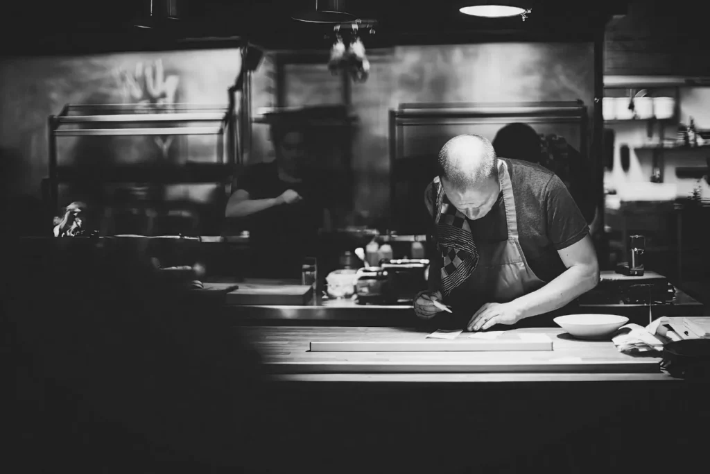 A high-contrast black and white photo of a bald chef leaning over a wooden counter, meticulously plating or preparing a dish. The background is a professional kitchen with industrial equipment.