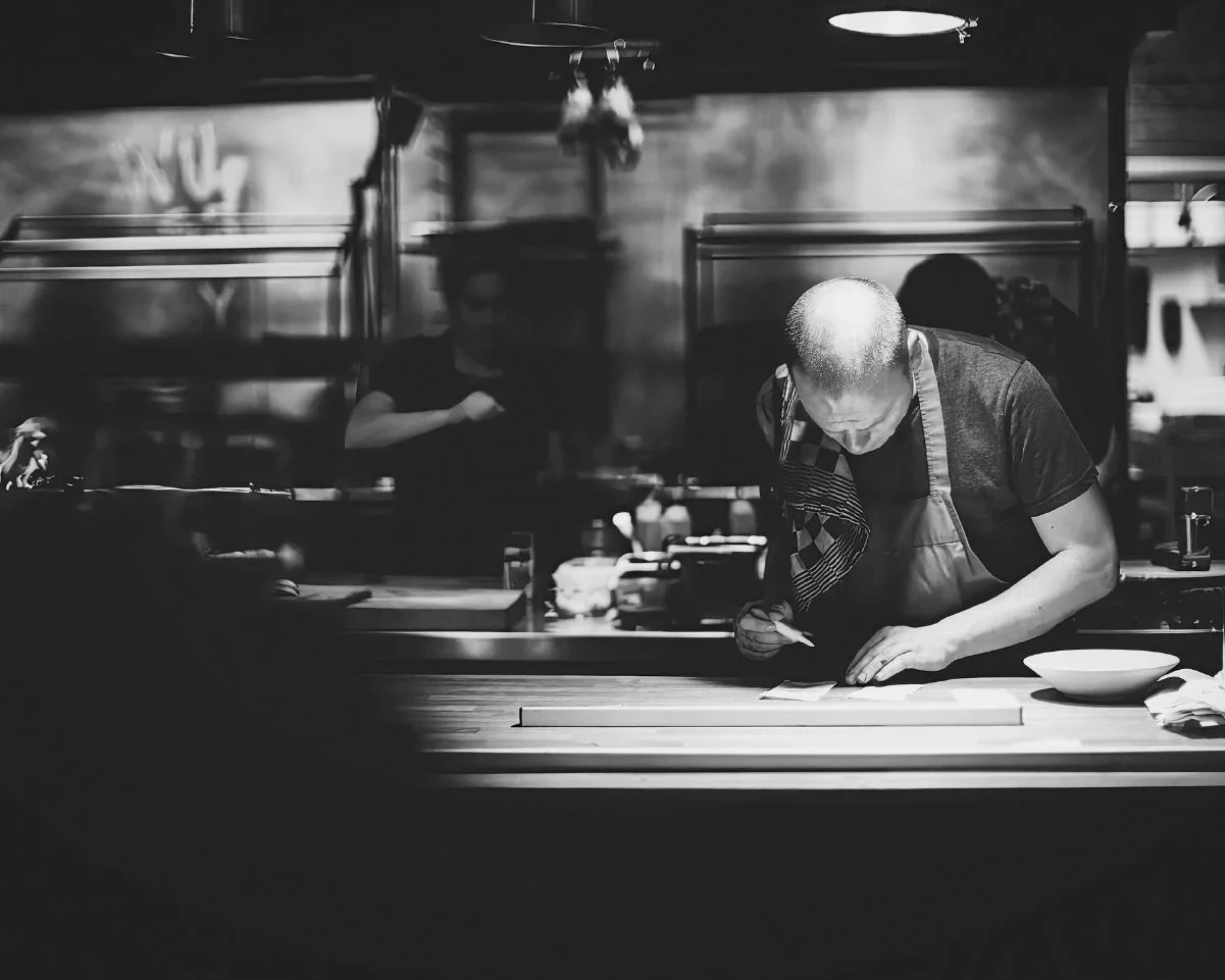 High-contrast black and white photograph of a bald chef intensely focused on plating a dish using tweezers, capturing the pressure, precision, and romanticized perfectionism often linked to toxic kitchen culture.