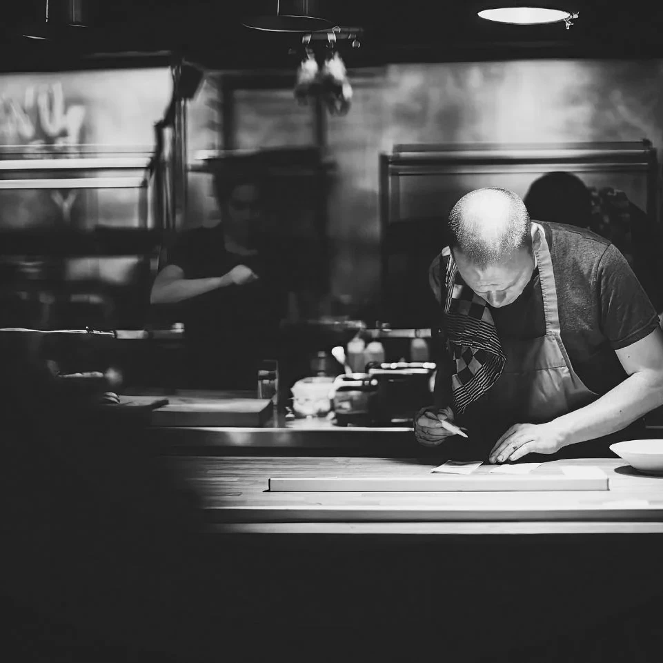 A high-contrast black and white photo of a bald chef leaning over a wooden counter, meticulously plating or preparing a dish. The background is a professional kitchen with industrial equipment.