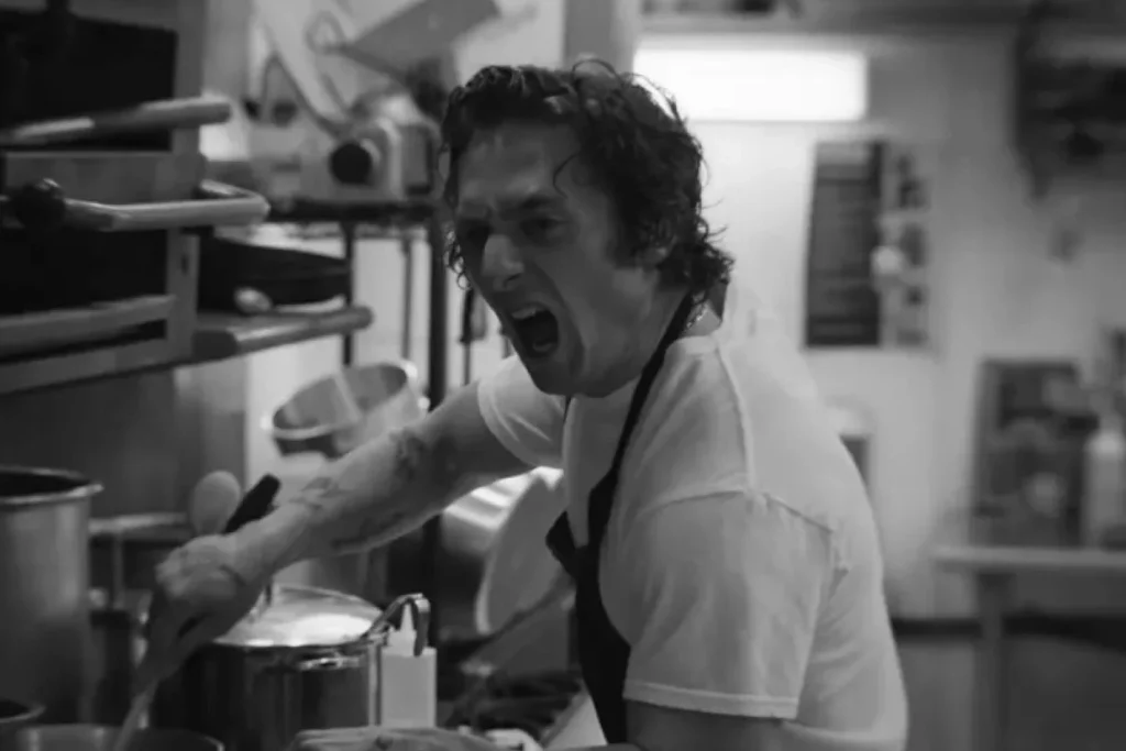 A black and white close-up of a chef with tattoos on his arm, shouting or expressing intense frustration while working over a stove in a cluttered, busy kitchen.