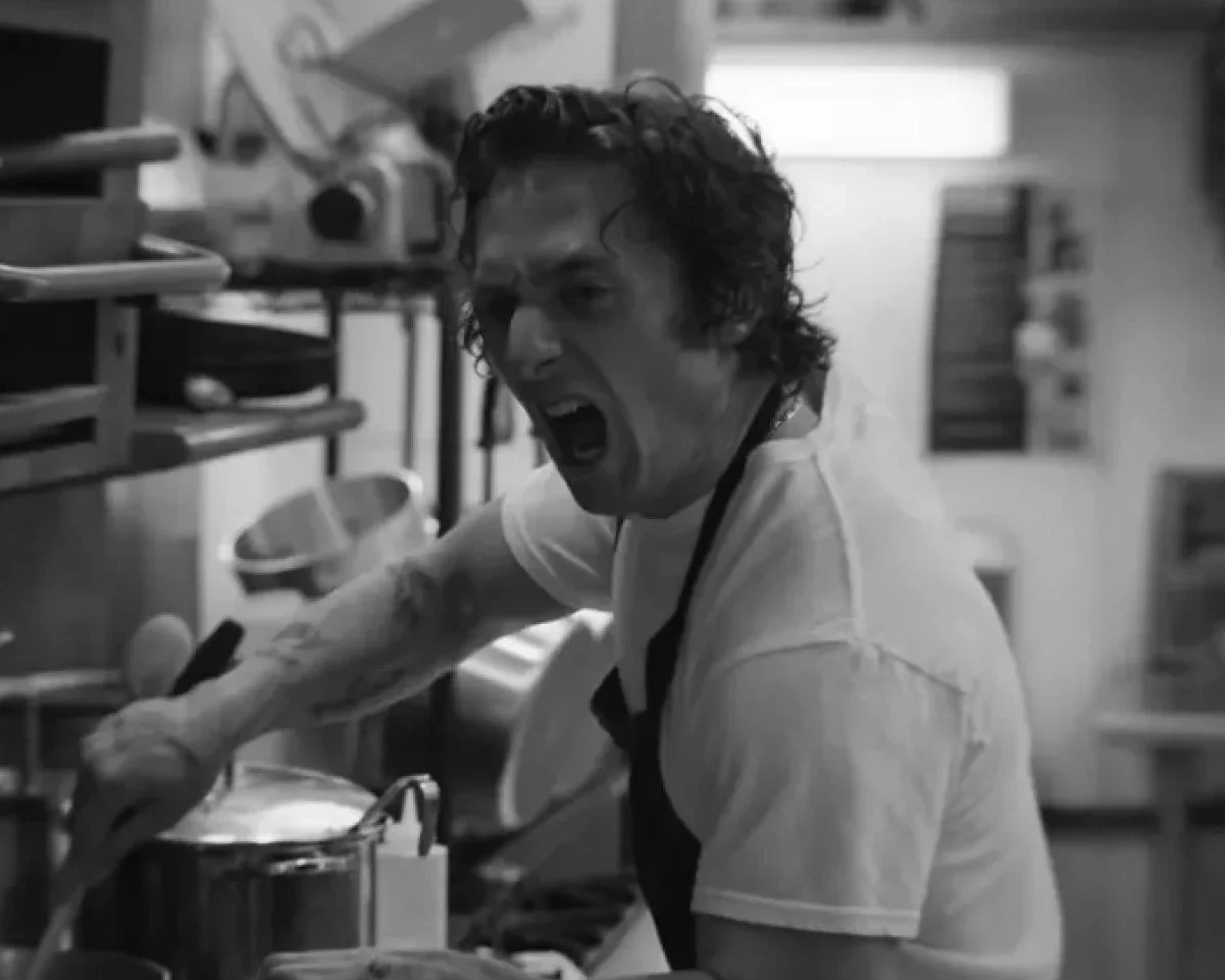 A black and white close-up of a chef with tattoos on his arm, shouting or expressing intense frustration while working over a stove in a cluttered, busy kitchen.